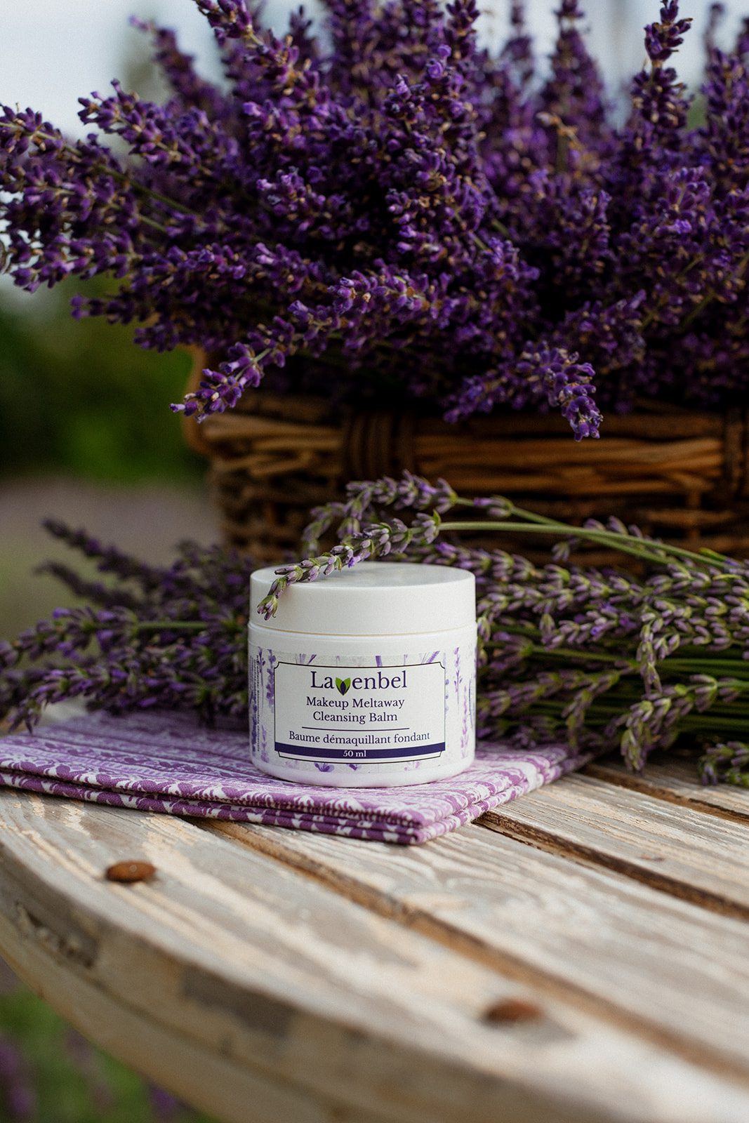 Melting cleansing balm (Lavenbel, 50 ml) on a table, with a lavender bouquet and basket in the background.